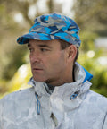 Man in a blue and gray camouflage cap and a light-colored Result RC59X jacket with blue accents, standing outdoors with greenery in the background.