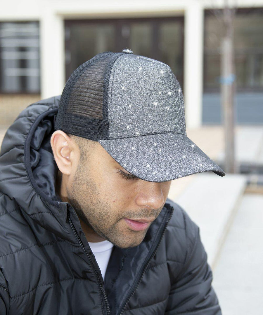 Man wearing a black puffer jacket and a sparkling black mesh cap with star-like designs, sitting outdoors, sporting the RC90X cap by Result.
