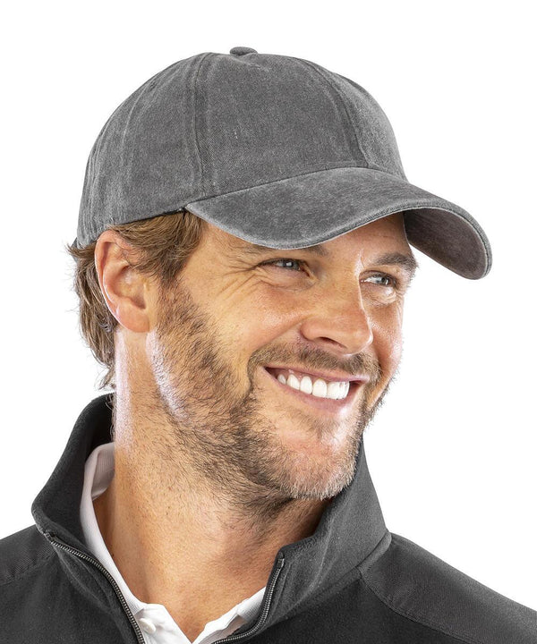A man with light brown hair and a short beard is smiling to the side, wearing the Result Headwear Core pure cotton vintage cap in dark grey and a black jacket over a white collared shirt against a plain white background.