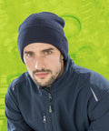 Man wearing a navy **Result RC933** beanie and blue jacket against a green background with circular patterns.