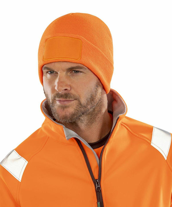 A person wearing an orange beanie and an Orange RC934 jacket with reflective strips from Result Genuine Recycled, looking off to the side.