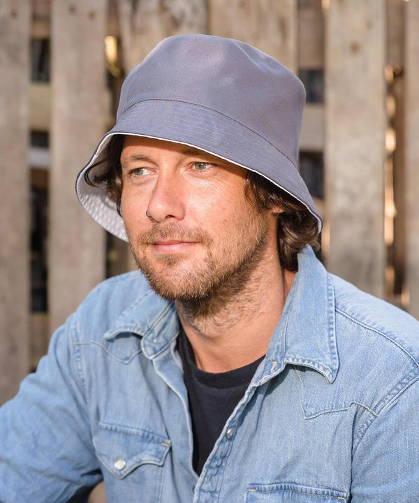A man with brown hair and a beard wears the Result Recycled Core recycled reversible bucket hat and a light blue denim shirt as he sits in front of a wooden fence, looking slightly to the side.