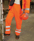 A person indoors wears Result Safeguard Safety high-vis trousers with reflective stripes, holds an orange safety helmet, and rests one foot on a metal ladder.