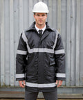 Man in a hard hat and reflective work jacket stands in front of a metal structure, looking directly at the camera. He is dressed in formal wear underneath the jacket, including a tie and trousers, sporting the Result RE23A.