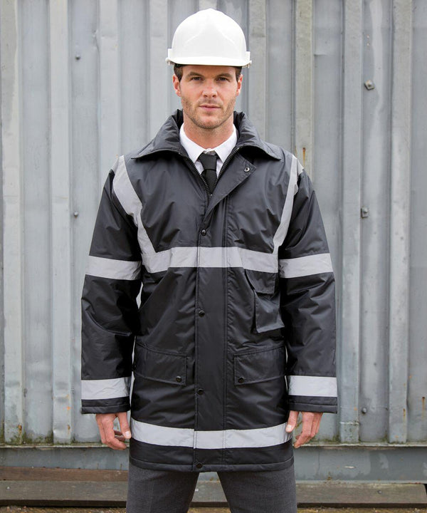 Man in a hard hat and reflective work jacket stands in front of a metal structure, looking directly at the camera. He is dressed in formal wear underneath the jacket, including a tie and trousers, sporting the Result RE23A.