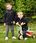 Two children, one standing and the other sitting on a tricycle, are outdoors on a grassy area with foliage in the background. Both are wearing Result RE33J black jackets and sneakers.