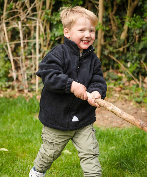 Young child wearing a Result RE36J black zip-up jacket and khaki pants holds a large stick while smiling outdoors.