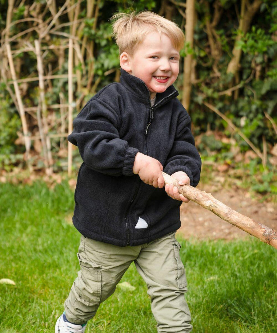 Young child wearing a Result RE36J black zip-up jacket and khaki pants holds a large stick while smiling outdoors.