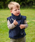 A young child stands on grass holding a tent peg in one hand, wearing a Result RE37J navy sleeveless jacket over a gray long-sleeved shirt.