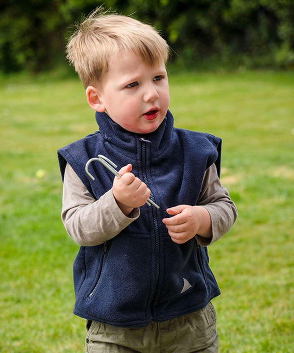 A young child stands on grass holding a tent peg in one hand, wearing a Result RE37J navy sleeveless jacket over a gray long-sleeved shirt.