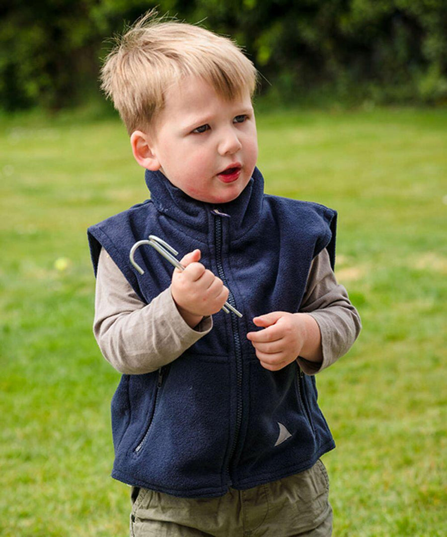 A young child stands on grass holding a tent peg in one hand, wearing a Result RE37J navy sleeveless jacket over a gray long-sleeved shirt.
