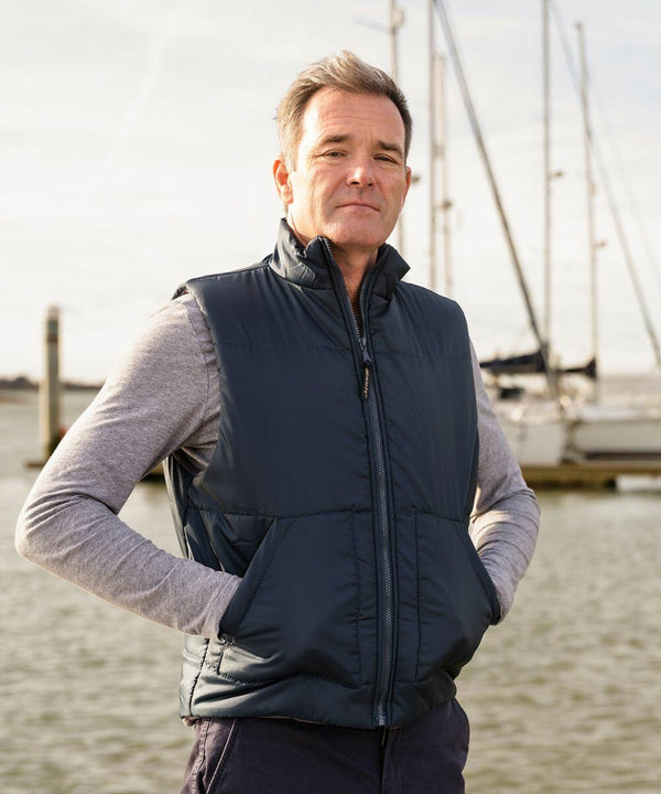 A man stands with hands in pockets, wearing a Result RE44A navy vest and grey long-sleeve shirt, in front of a marina with sailboats.