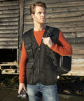 A man in an orange sweater and black vest carrying a bag over his shoulder stands in front of a weathered wooden building, holding a Result RE45A in his left hand.