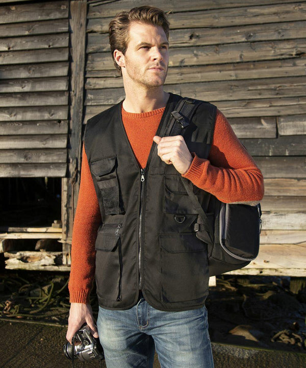 A man in an orange sweater and black vest carrying a bag over his shoulder stands in front of a weathered wooden building, holding a Result RE45A in his left hand.