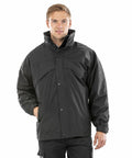A person wearing a black Result RE68A zip-up jacket with a high collar, standing with a neutral expression and one hand in a pocket.