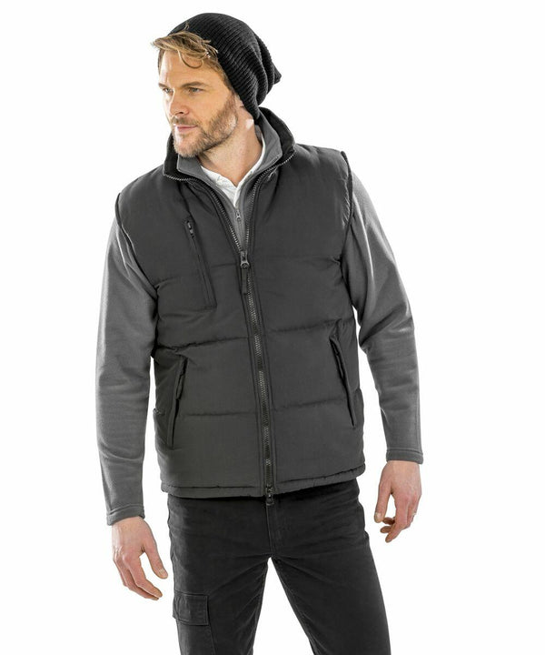 A man with a beard wearing a black beanie, a Result RE88A gray zip-up vest, a gray long sleeve shirt, and black pants stands against a white background.