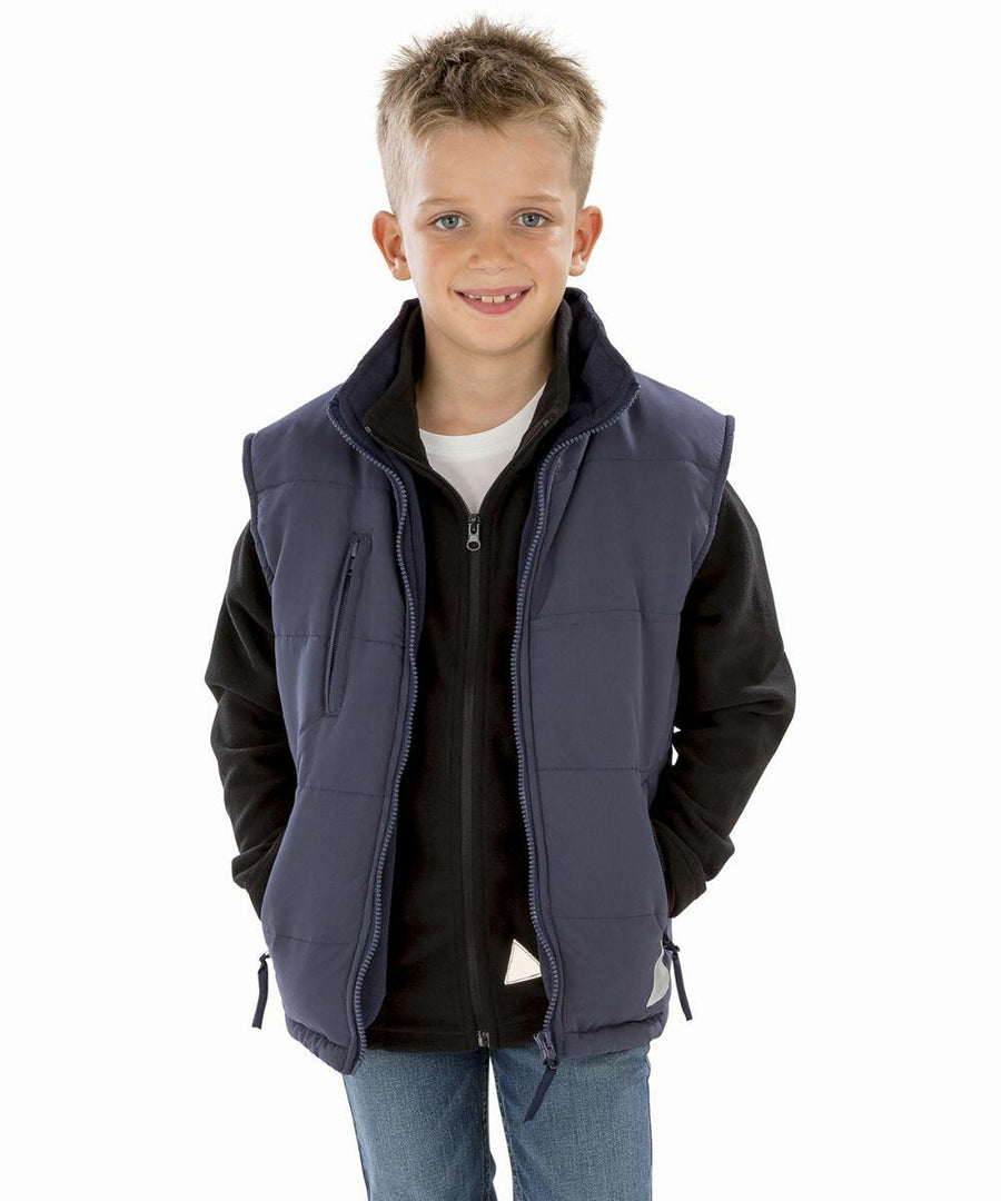 A young boy with short, light hair is wearing a black jacket and a navy blue sleeveless vest. He stands smiling with his hands in his pants pockets against a white background, wearing the Result RE88J.