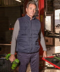 A man wearing a padded vest and long-sleeve shirt holds a green Result RE94A in a workshop with equipment and machinery visible in the background.