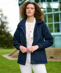 A person with curly hair stands outdoors, wearing a navy blue Regatta RG052 over a white shirt and pants. They are in front of a structure with glass windows and green framing.