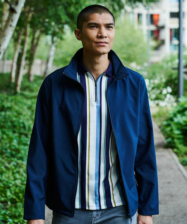 A man wearing a Regatta RG073 over a striped shirt stands outdoors on a path with greenery and buildings in the background.