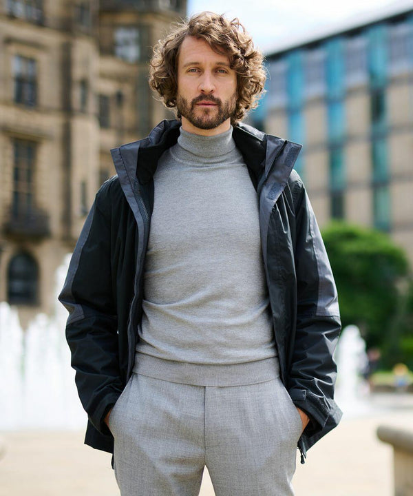 A man with curly hair wearing a grey RG085 sweater from Regatta and a black jacket stands outdoors in front of a building and a fountain.