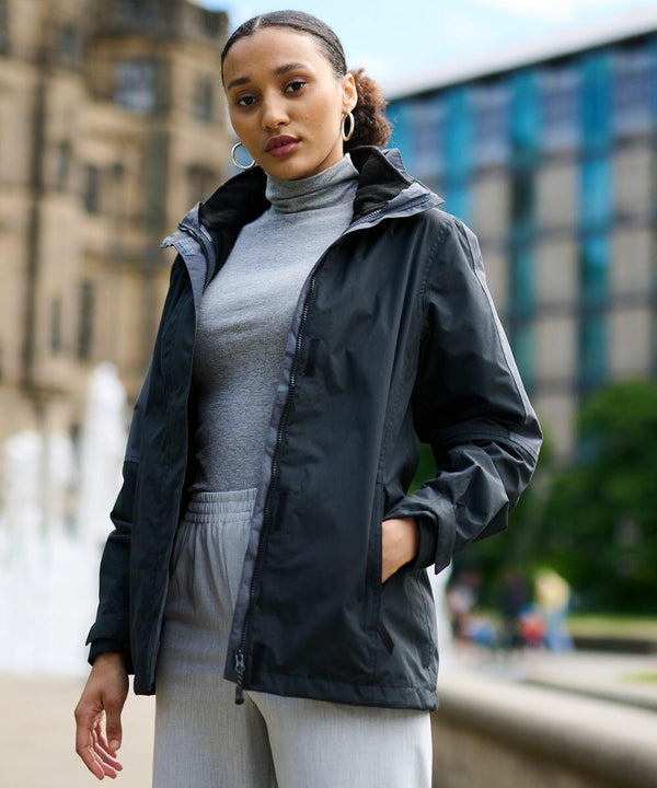 A person standing outdoors wearing a grey turtleneck, grey pants, and a black Regatta RG086 jacket, with one hand in their pocket. A fountain and buildings are visible in the background.