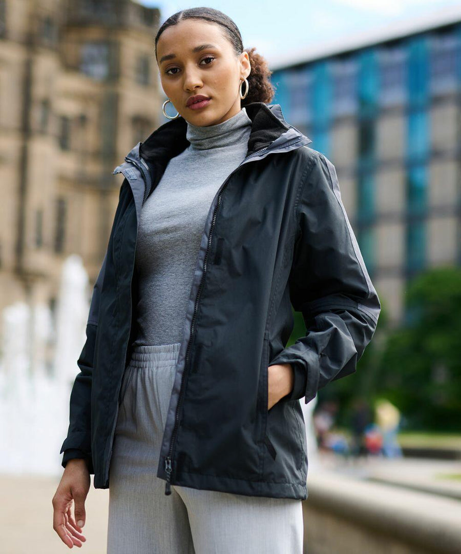 A person standing outdoors wearing a grey turtleneck, grey pants, and a black Regatta RG086 jacket, with one hand in their pocket. A fountain and buildings are visible in the background.
