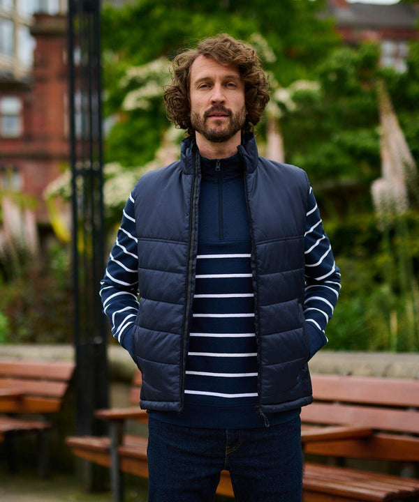 A man with curly hair stands outdoors wearing a navy Regatta Professional RG113 puffer vest over a striped shirt, with his hands in his pockets. Benches and greenery are visible in the background.