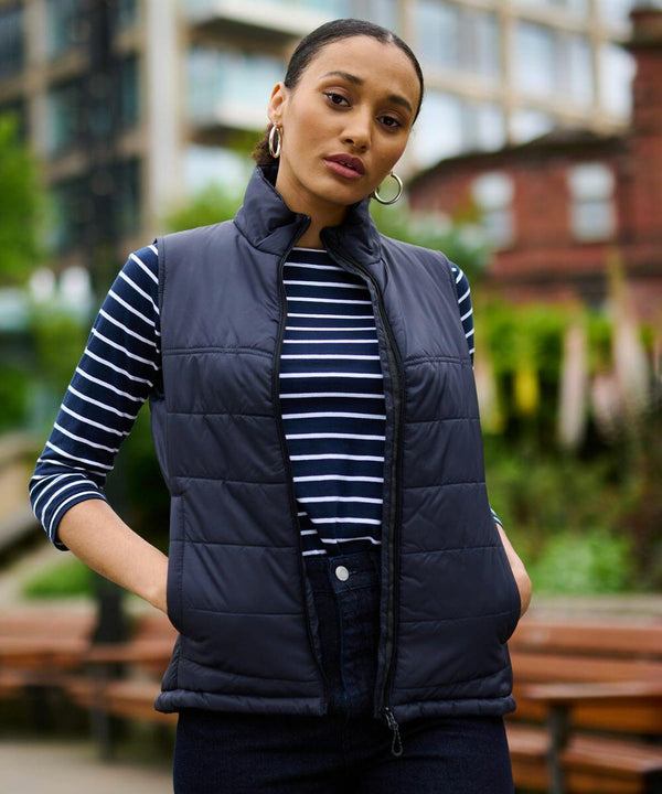 A person stands outdoors wearing a black Regatta RG114 over a striped shirt, with hands in pockets. Background includes buildings and greenery.