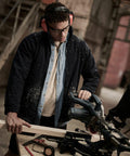 A person wearing safety goggles and earmuffs operates a Regatta RG122, cutting a piece of wood. Wood shavings are visible on their clothing. Various tools are scattered on the workbench.