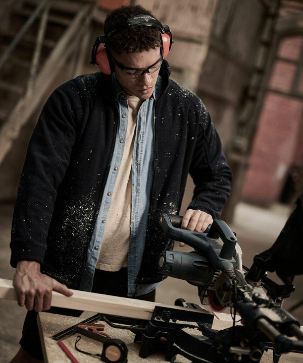 A person wearing safety goggles and earmuffs operates a Regatta RG122, cutting a piece of wood. Wood shavings are visible on their clothing. Various tools are scattered on the workbench.
