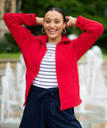 A person wearing a red jacket and striped shirt smiles with hands resting on the back of their head, standing in front of a fountain, showcasing the RG123 by Regatta.