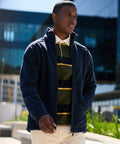 A man wearing a Regatta RG128 navy jacket walks outdoors. He has a striped shirt underneath and beige pants. The background includes a modern glass building.