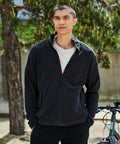 A person wearing a black Regatta RG131 zip-up jacket stands outdoors, hands in pockets, in front of a tree and a wall. A bicycle is partially visible next to them.