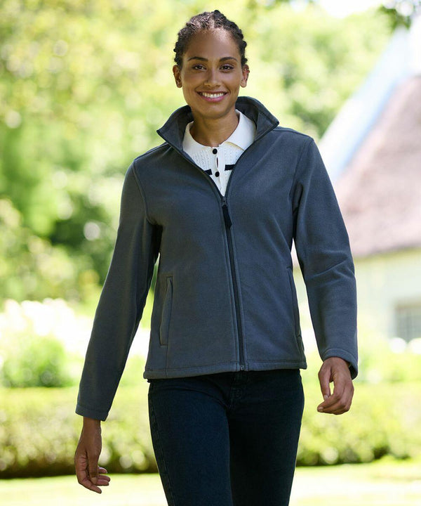 A person walks outdoors in a grey RG140 jacket by Regatta and dark jeans, smiling. The background is blurry, with greenery and a building visible.