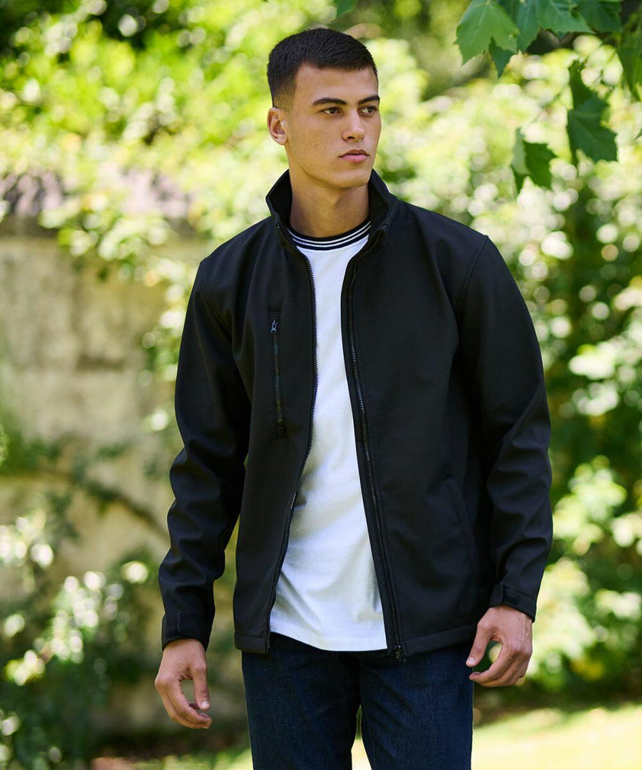 A man stands outdoors wearing a black Regatta RG147 zip-up jacket over a white shirt and blue jeans, with greenery in the background.