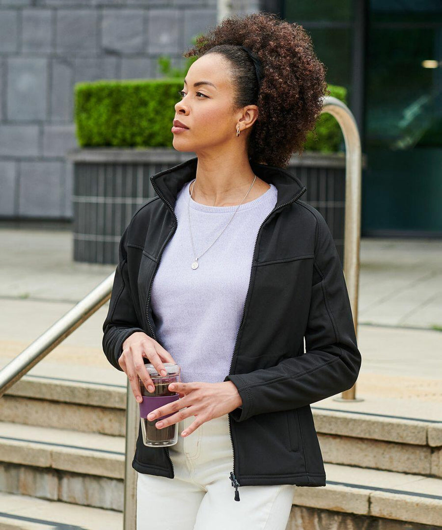 A person in a black jacket and white pants holds a glass of water while standing outside near stairs and greenery, wearing RG151 by Regatta.