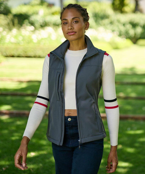 A woman walks in a park wearing a grey Regatta RG155 vest over a white long-sleeve shirt with striped armbands. She has braided hair and is outdoors on a sunny day.