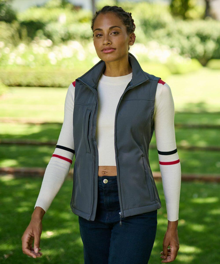 A woman walks in a park wearing a grey Regatta RG155 vest over a white long-sleeve shirt with striped armbands. She has braided hair and is outdoors on a sunny day.