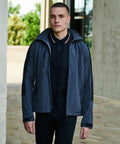 A person is standing outdoors wearing a dark jacket over a black polo shirt, identified as the RG157 by Regatta. The background shows a concrete structure and greenery.