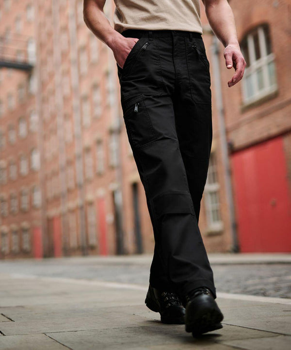 Person walking down a street lined with brick buildings, wearing Regatta RG171 black cargo pants, a beige shirt, and black shoes.
