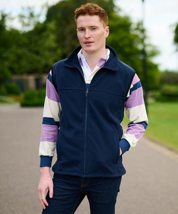 A person with red hair is standing outdoors, wearing a navy Regatta RG182 vest over a striped shirt with pink, white, and blue sleeves, and blue jeans. Trees and a pathway are in the background.