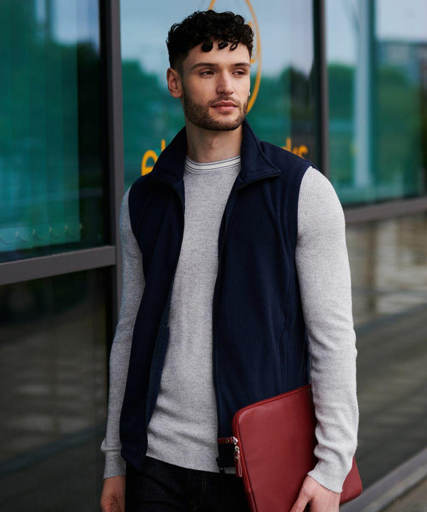 Man with short curly hair wearing a navy vest over a gray sweater holds a Regatta RG185 laptop case while standing outside a glass building.