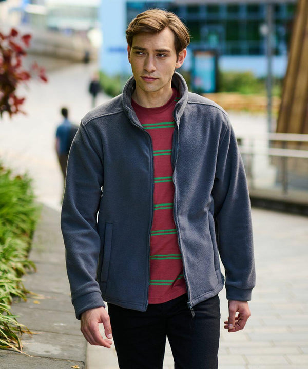 A person wearing a grey Regatta RG189 jacket and red striped shirt walks outdoors near a path with plants and buildings in the background.