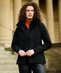 A person with curly hair is standing outdoors wearing a black jacket and an orange turtleneck, with a building featuring columns in the background. They are holding the Regatta RG192.