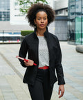 A person with curly hair, wearing a black jacket and polka-dot top, walks outside holding a red RG195 folder by Regatta. Urban buildings are in the background.