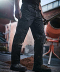 A worker wearing dusty black RG233 cargo pants and boots from Regatta stands holding a shovel, with a concrete mixer and a pile of sand in the background.