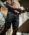 A person in a brown shirt and black work shorts is holding a Regatta RG237, standing next to a wooden surface in a carpentry or construction setting.