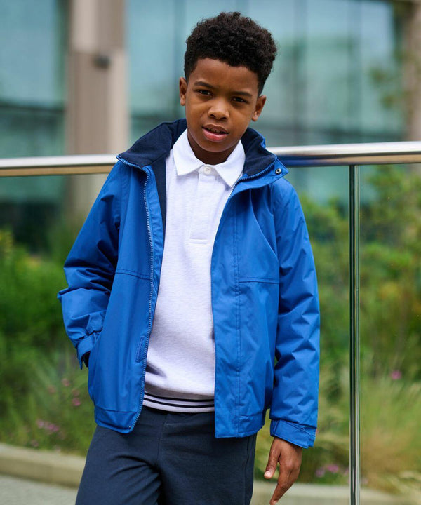A young boy in a Regatta RG244 blue jacket and white polo shirt stands outdoors near a glass railing.
