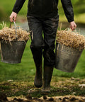 Person carrying two RG254 buckets filled with straw while walking on grass, dressed in black Regatta rain gear and boots.